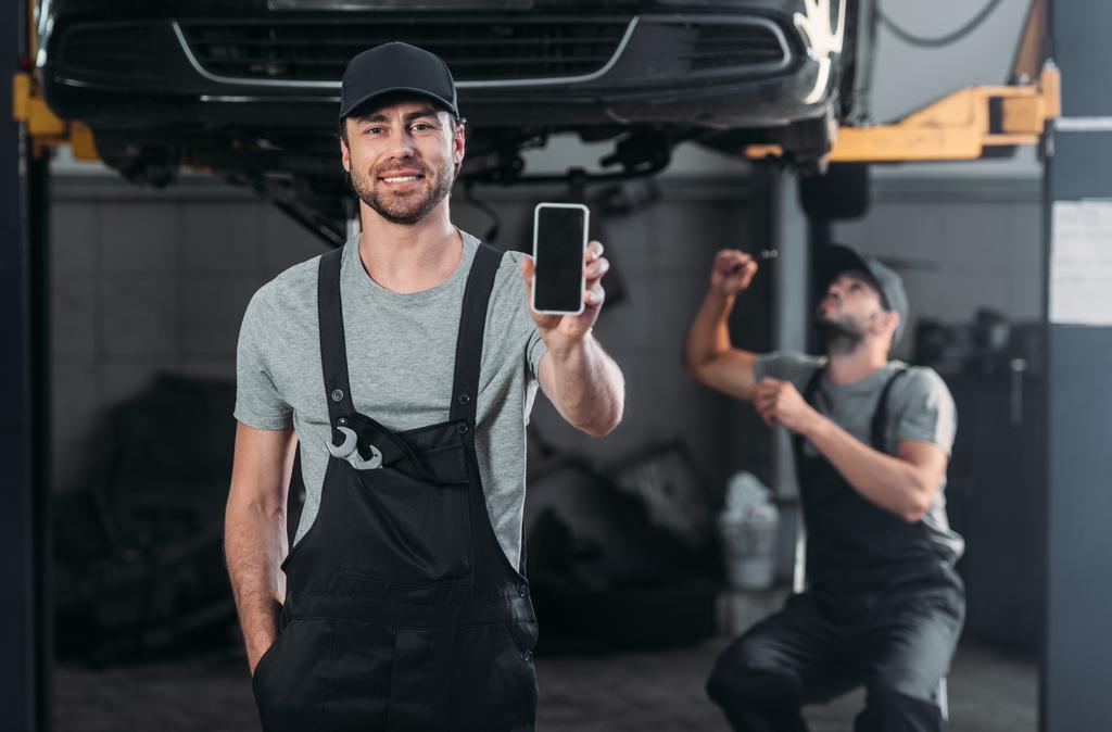 auto mechanik představí smartphone s prázdnou obrazovkou, zatímco kolega pracující v dílně za - Fotografie, Obrázek