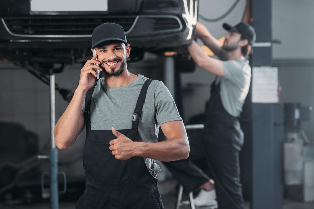 auto mechanik hovoří o smartphone a ukazuje palec nahoru, zatímco kolega pracující v dílně za - Fotografie, Obrázek