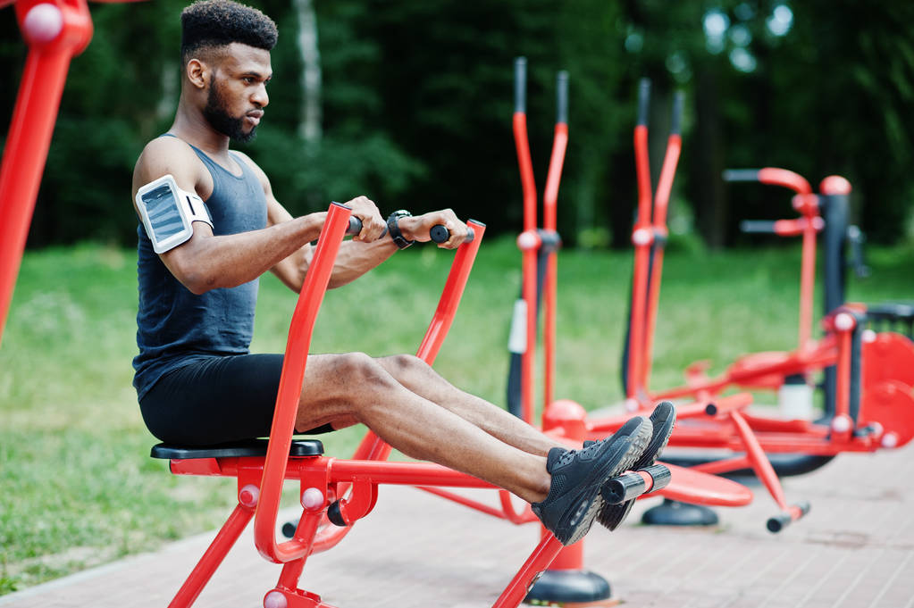Hombre atleta afroamericano con funda de brazo deportivo para teléfono móvil, haciendo ejercicios en los simuladores de entrenamiento callejeros
. - Foto, Imagen