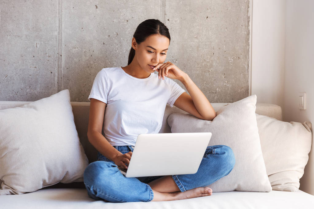 Pensivo jovem asiático mulher usando laptop computador enquanto sentado em um sofá em casa
 - Foto, Imagem