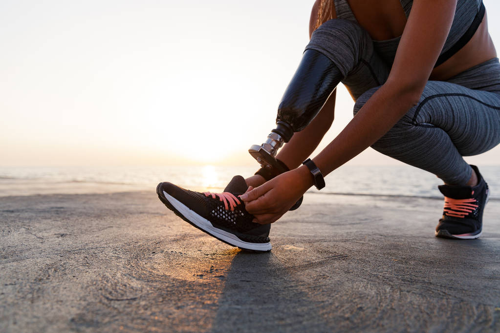 Close up of disabled athlete woman with prosthetic leg tying her shoelace outdoor at the beach - Photo, Image