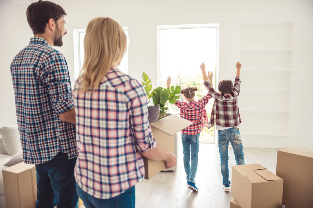 Mladí šťastné rodiny čtyři osoby, rodiče drží boxy krabička s věci věci v obýváku světle stěhování do nového bytu, nedospělý děti hrají - Fotografie, Obrázek