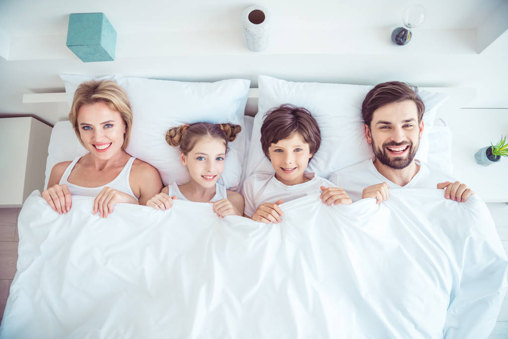 Młody szczęśliwy uśmiechający się rodziny czterech osób razem leżącego w łóżku śmiejąc się pokryte prześcieradła koc. Góry powyżej wysoki kąt widzenia - Zdjęcie, obraz