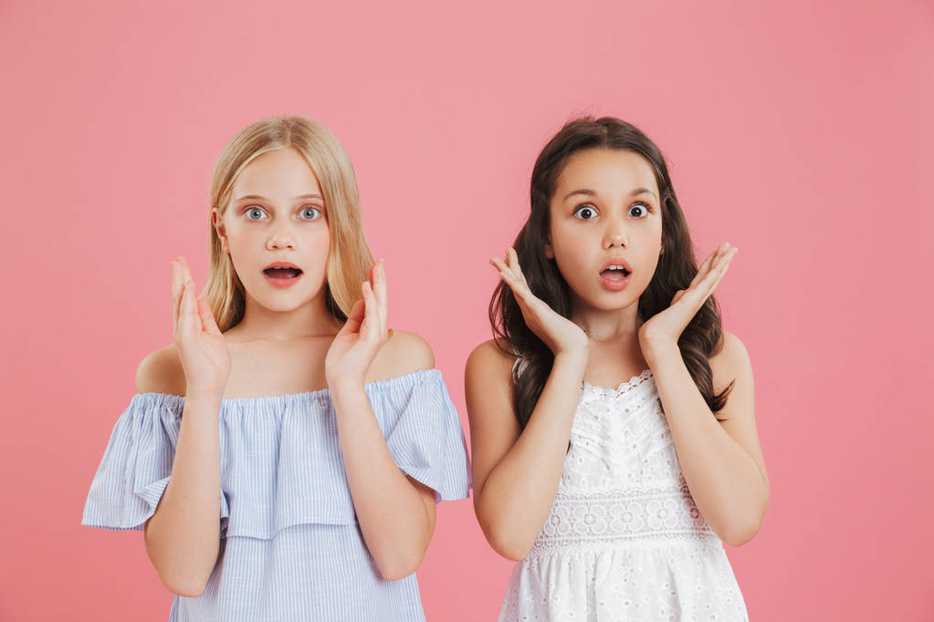 Foto van Europese kleine meisjes het dragen van jurken uiten verrassing en verhogen handen gezicht met open mond geïsoleerd op roze achtergrond - Foto, afbeelding