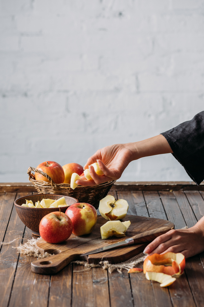 tiro cortado de mulher com pedaços de maçã na mão e maçãs frescas na tábua de corte em mesa de madeira
 - Foto, Imagem