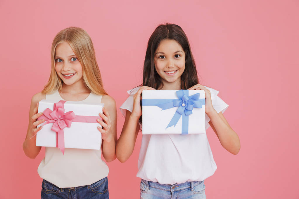 Two adorable caucasian girls 8-10 in casual clothing smiling at camera and holding present boxes with colorful bows isolated over pink background - Photo, Image