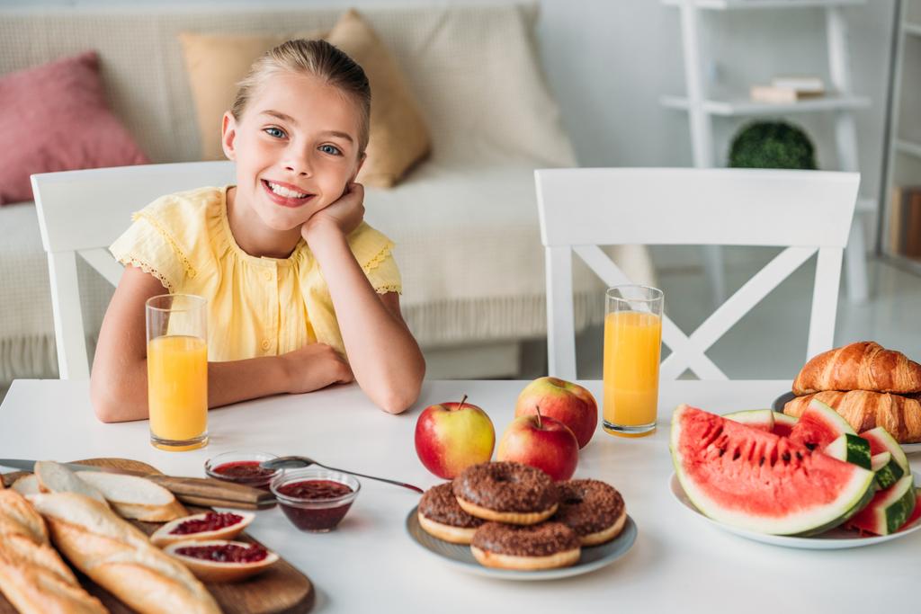 Imádnivaló kis gyerek otthon reggelire különböző élelemmel asztalnál ül - Fotó, kép