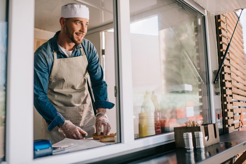 guapo sonriente joven chef masculino mirando hacia otro lado mientras cocina en camión de comida
 - Foto, imagen