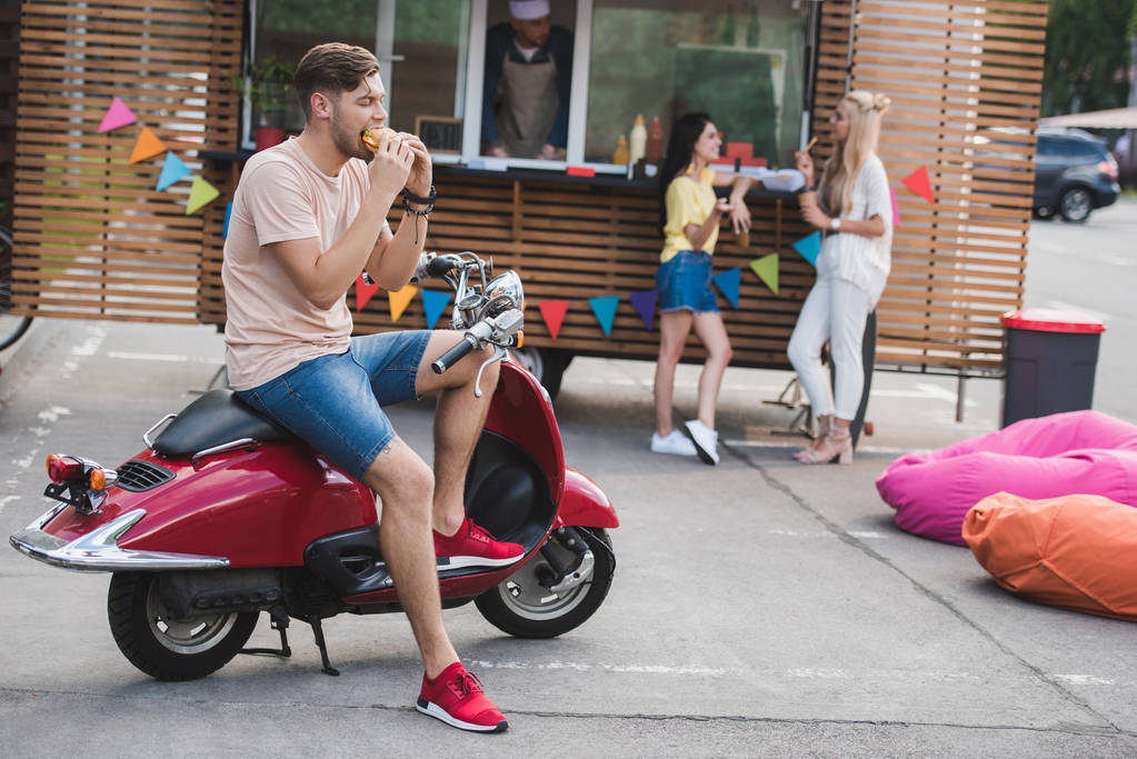 Mann isst Burger und sitzt auf Motorrad in der Nähe von Foodtruck - Foto, Bild
