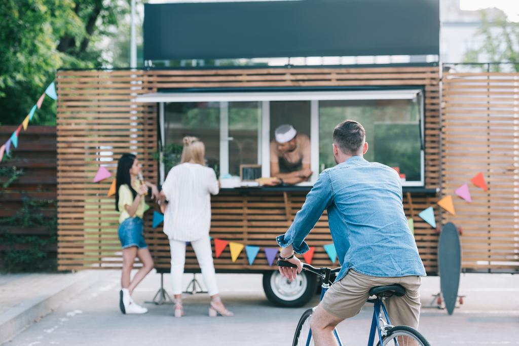 Rückansicht eines Mannes, der in der Nähe eines Foodtrucks auf einem Fahrrad sitzt - Foto, Bild