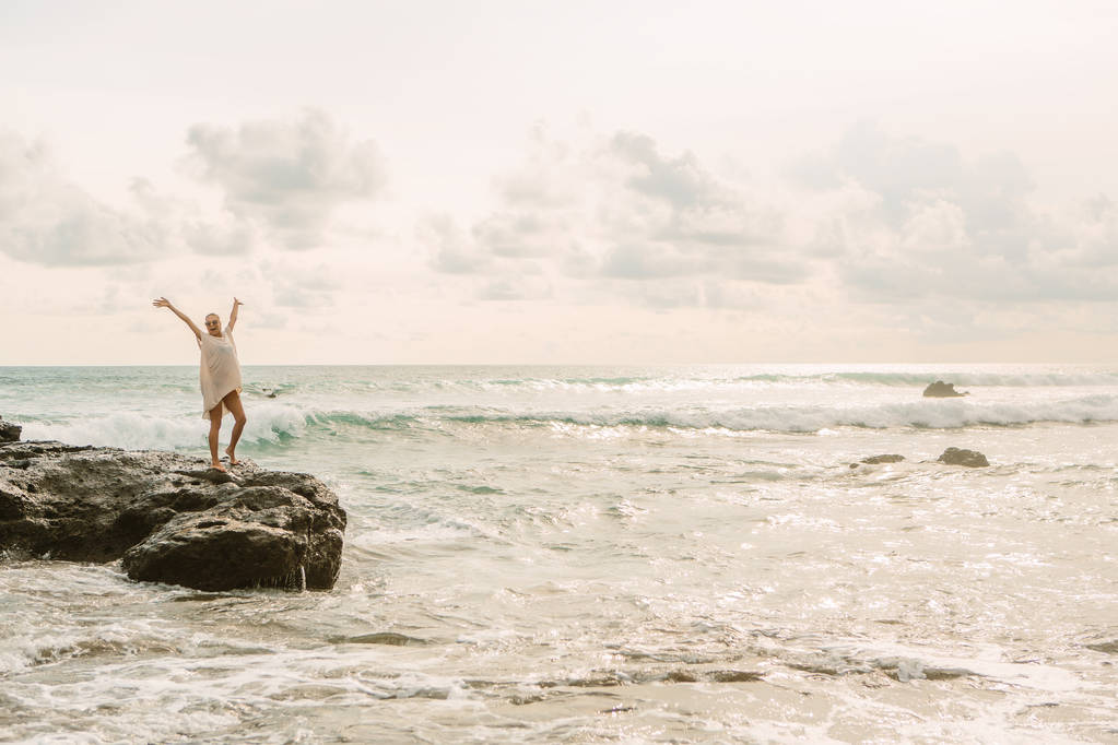 Mooi meisje glimlach op camera tijdens verblijf op steen voor grote golven op de rotsen. Sexy dame op strand zee zonsondergang of Oceaan zonsopgang. Gevaar, waarschuwing, reizen en actieve levensstijl concept. - Foto, afbeelding