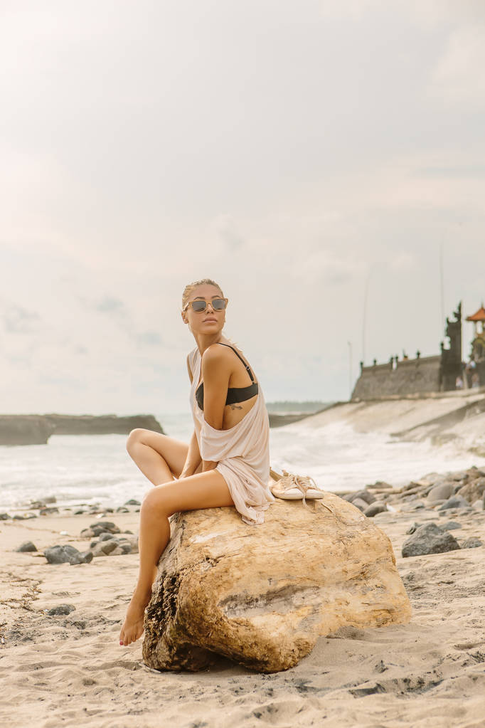 女性の自然の美しさ。大きな石の上にかわいい女の子が座っています。夕日や海の日の出を楽しむ海辺のエキゾチックな白砂のビーチで若い女性。旅行、サーファーのリラックスやアクティブなヨガ瞑想ライフ スタイルのコンセプト. - 写真・画像