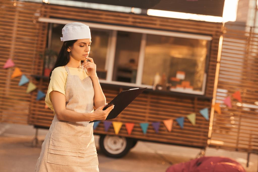 attractive pensive chef in apron holding clipboard near food truck - Photo, Image