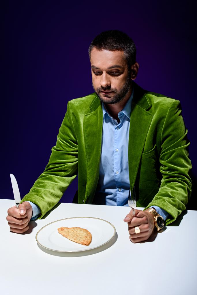 человек в стильной зеленой бархатной куртке сидит за столом с мясной выпечкой на тарелке с синим фоном позади
 - Фото, изображение
