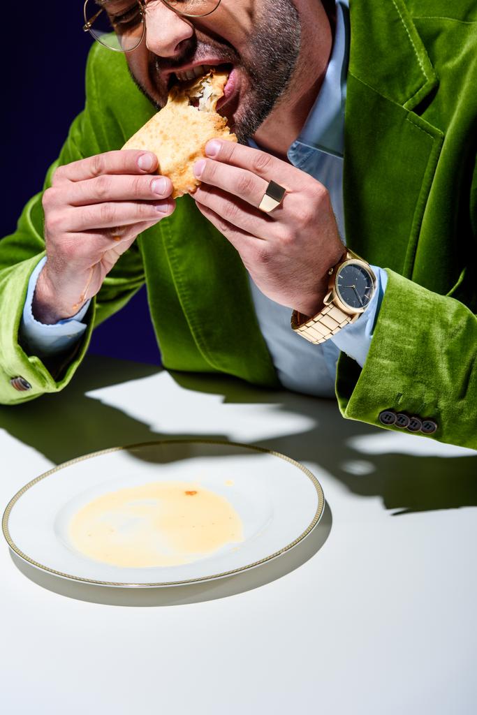 levágott lövés ember eszik cheburek, kék háttér mögött asztalnál elegáns zöld bársony kabát - Fotó, kép