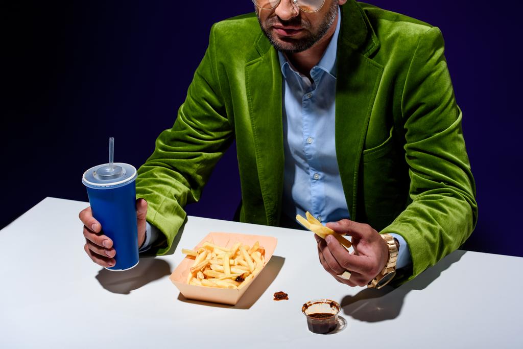 részleges Nézd divatos ruházatot és kólát inni, sült krumplival, és ketchup kék hátteret asztalnál ülő ember - Fotó, kép
