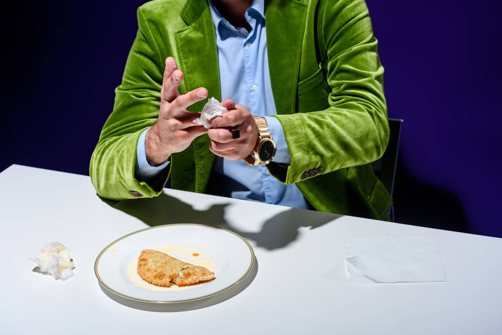 περικοπεί shot του ανθρώπου σε βελούδινο σακάκι ξήρανση τα χέρια με χαρτοπετσέτα, ενώ κάθεται στο τραπέζι με cheburek κρέας σε πιάτο με μπλε φόντο - Φωτογραφία, εικόνα