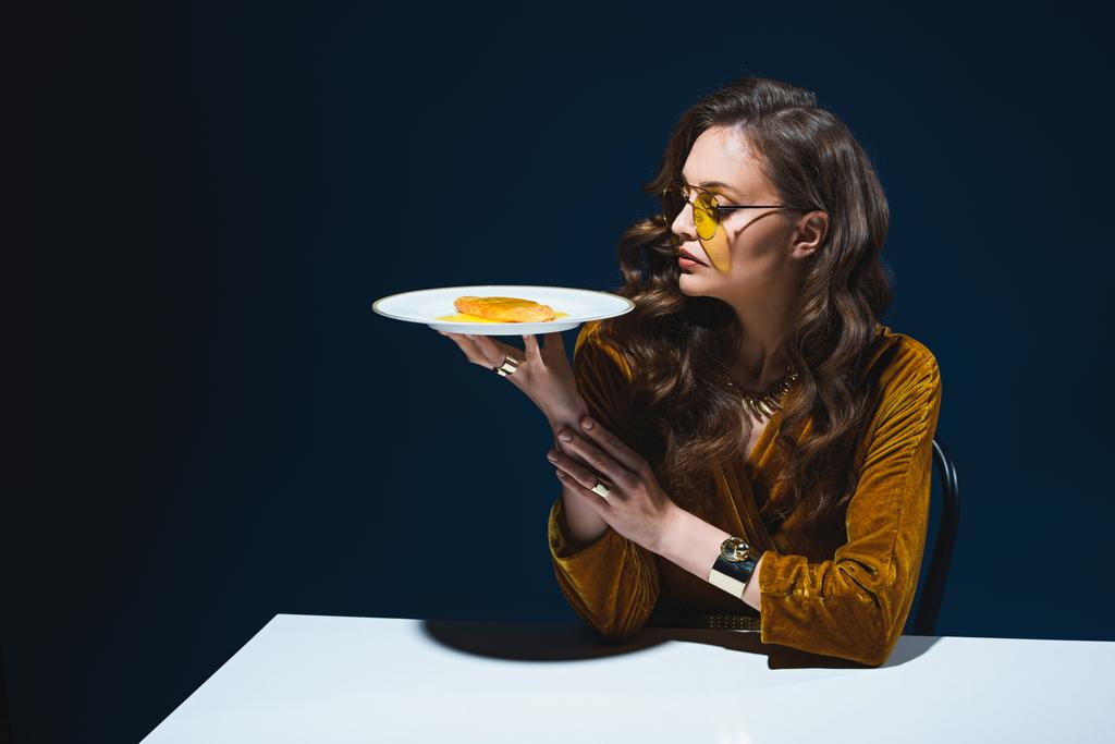 schöne Frau in stilvoller Kleidung mit ungesundem Fleischgebäck auf Teller sitzt am Tisch mit blauem Hintergrund hinter - Foto, Bild