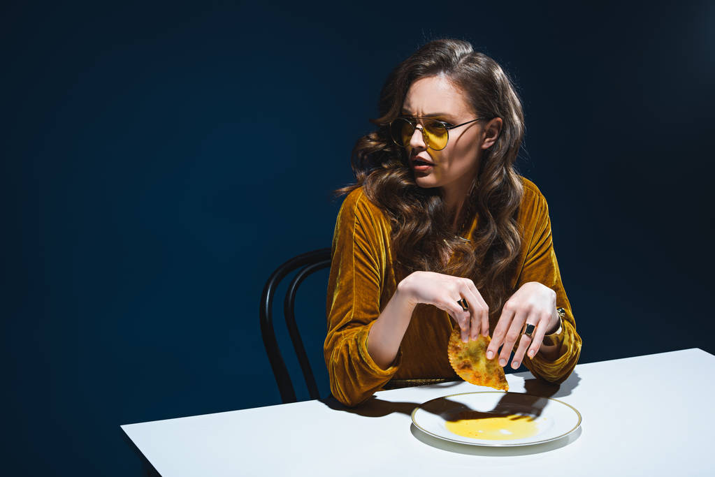 fashionable woman with unhealthy meat pastry sitting at table with blue backdrop - Photo, Image