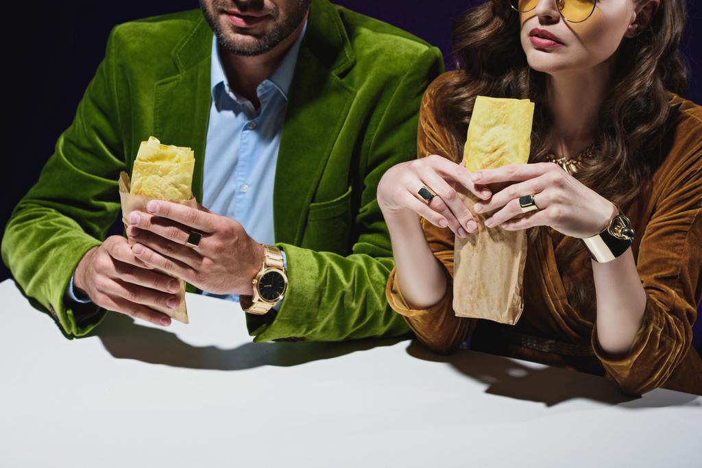 Teilansicht eines Paares in luxuriöser Samtkleidung mit Shawarma am Tisch sitzend mit schwarzem Hintergrund dahinter  - Foto, Bild