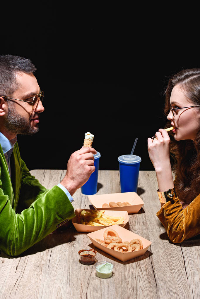 vista lateral de pareja en ropa de terciopelo elegante comiendo aros de cebolla frita, papas fritas y salsas en la mesa con fondo negro
 - Foto, imagen