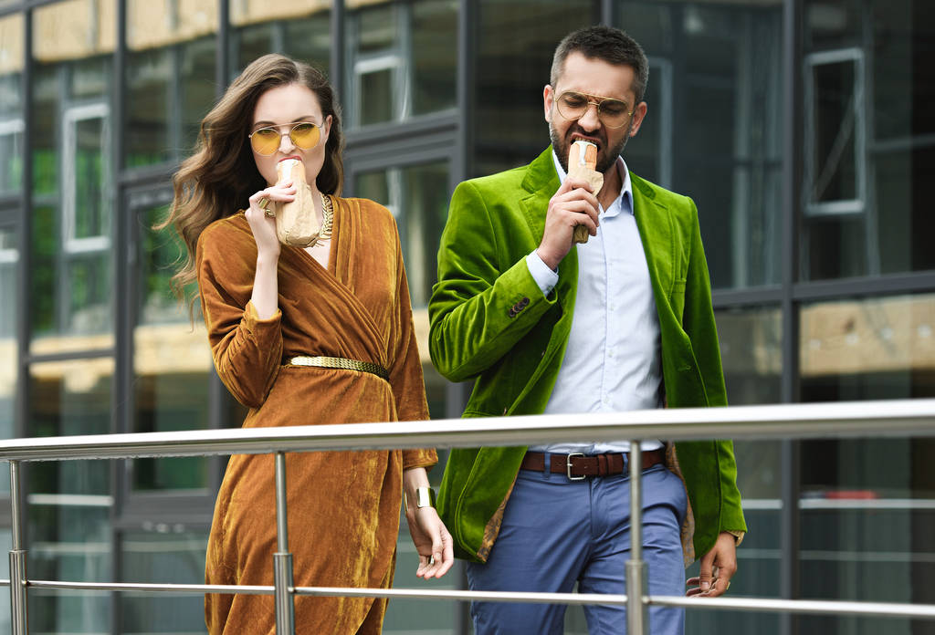 Portret van paar in luxe outfit eten hotdogs Frans tijdens het lopen op straat - Foto, afbeelding