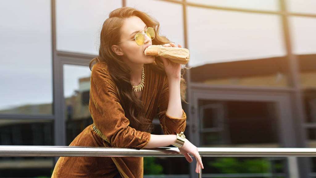 portré, vonzó nő, a cég luxus minőségű ruházatot és napszemüveg eszik francia hot-dog utcában - Fotó, kép