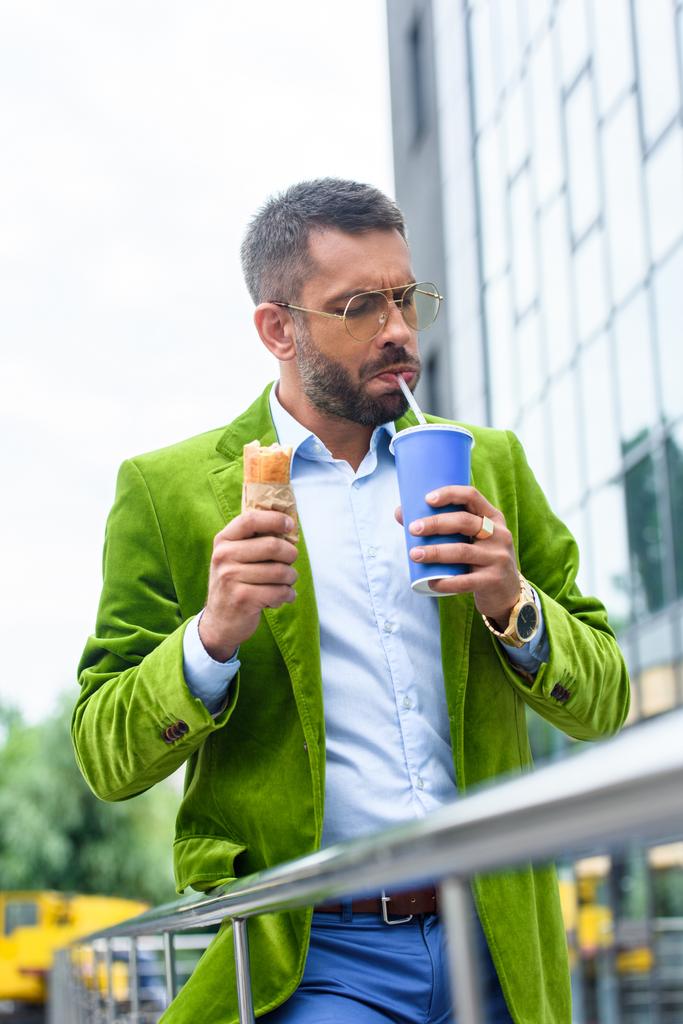 Porträt eines Mannes in grüner Samtjacke mit französischem Hot Dog, der auf der Straße Limonade trinkt - Foto, Bild