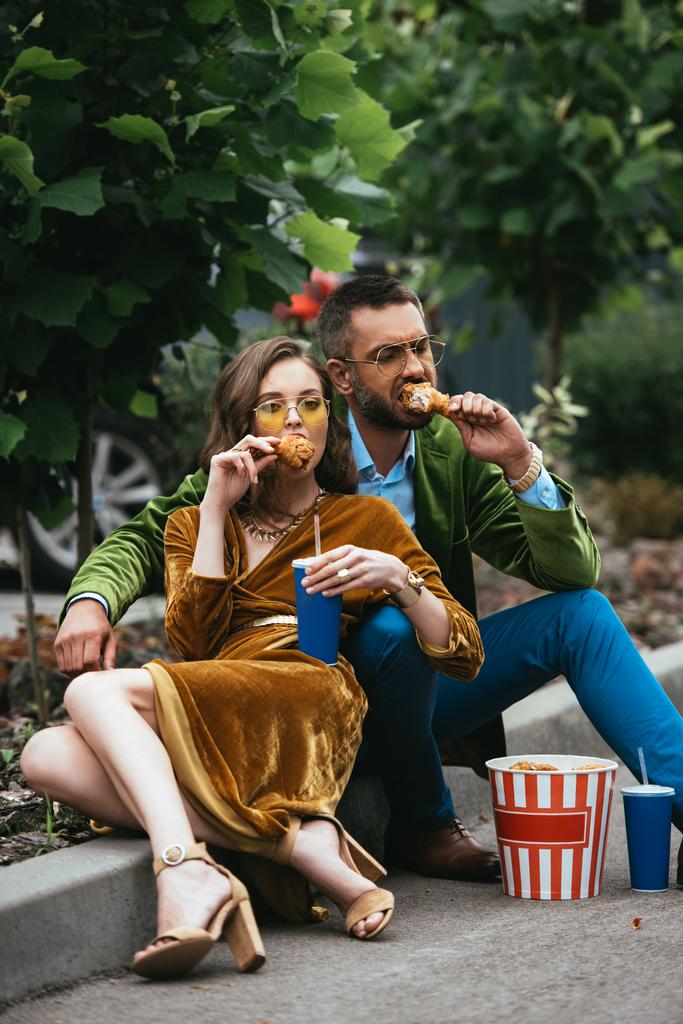 μοντέρνο ζευγάρι στο βελούδο ρούχα τρώει τηγανητό κοτόπουλο πόδια στον δρόμο - Φωτογραφία, εικόνα