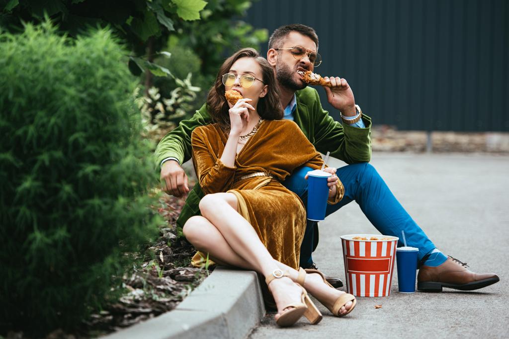 pareja de moda en ropa de terciopelo comiendo patas de pollo frito en la calle
 - Foto, imagen