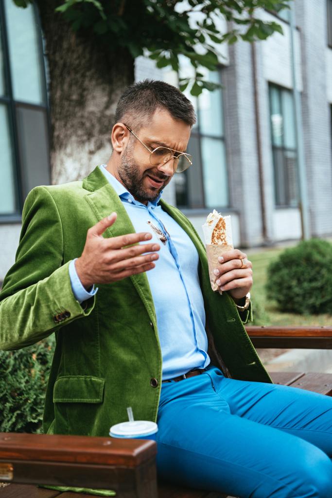 μοντέρνα άνθρωπος στο βρώμικο πουκάμισο με shawarma στο χέρι που κάθεται στον πάγκο στην οδό - Φωτογραφία, εικόνα
