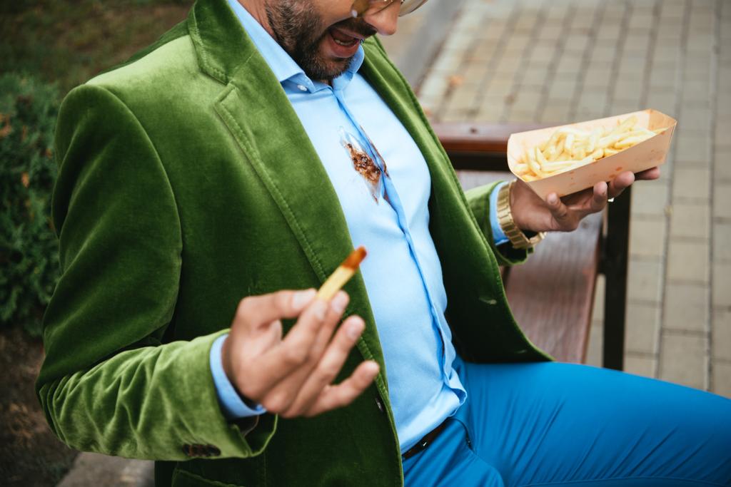 částečný pohled člověka v zelené Sametové sako s kečupem na košili a hranolky v rukou sedí na lavičce na ulici - Fotografie, Obrázek