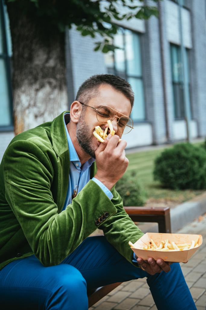 portrait of man in green velvet jacket eating french fries while sitting on bench on street - Photo, Image