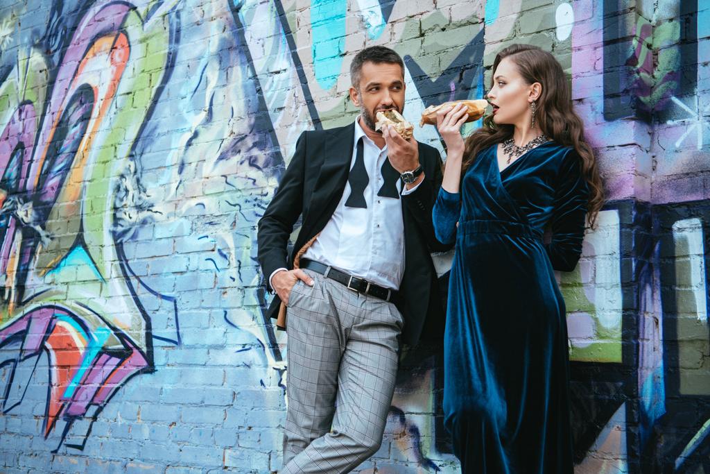couple in luxury clothing eating hot dogs near wall with graffiti on street - Photo, Image