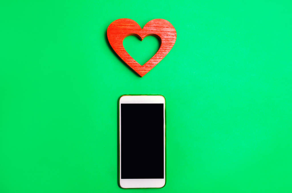 Abhängigkeit von sozialen Netzwerken. Smartphone und Herz auf grünem Hintergrund. Online-Dating, Flirten, Nachrichten und Telefonate mit dem Liebsten. Konzept der Liebe und Kommunikation - Foto, Bild