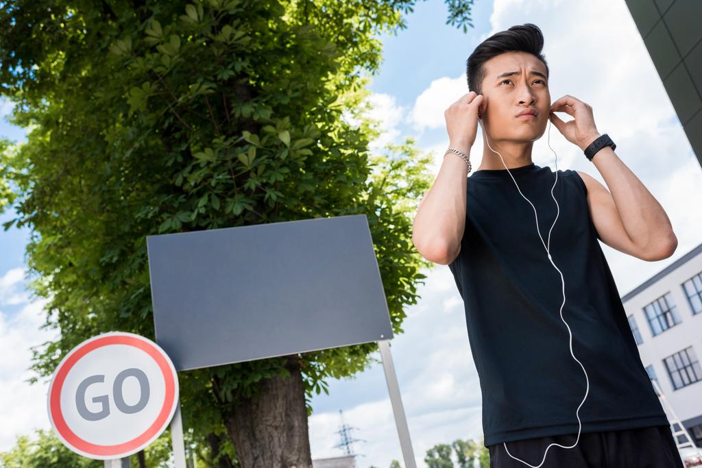low angle view of serious asian sportsman putting on earphones at street with empty signboard and road sign with lettering go - Photo, Image
