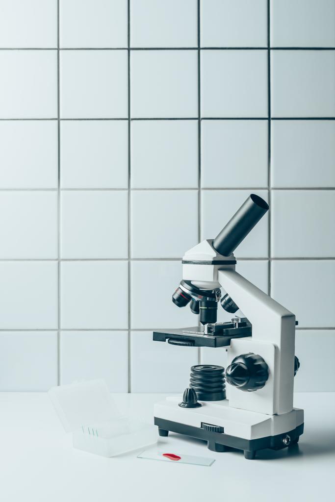 Blutprobe und Lichtmikroskop auf weißer Tablette vor gekachelter Wand - Foto, Bild