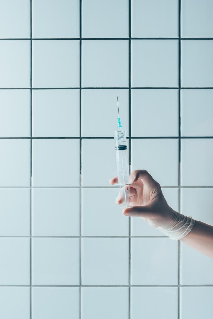Schnappschuss eines Arztes in Handschuhhalterspritze mit transparenter Flüssigkeit vor gekachelter weißer Wand - Foto, Bild