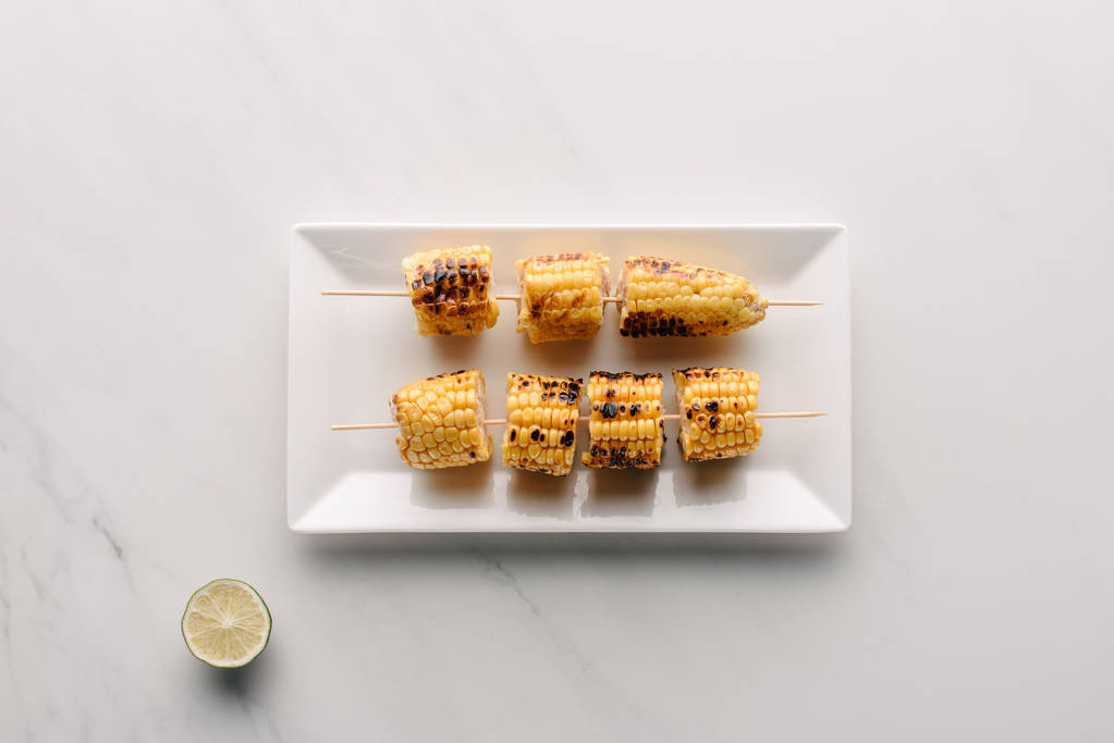 bovenaanzicht van gegrild maïs op plaat in de buurt van kalk slice op marmeren tafel  - Foto, afbeelding