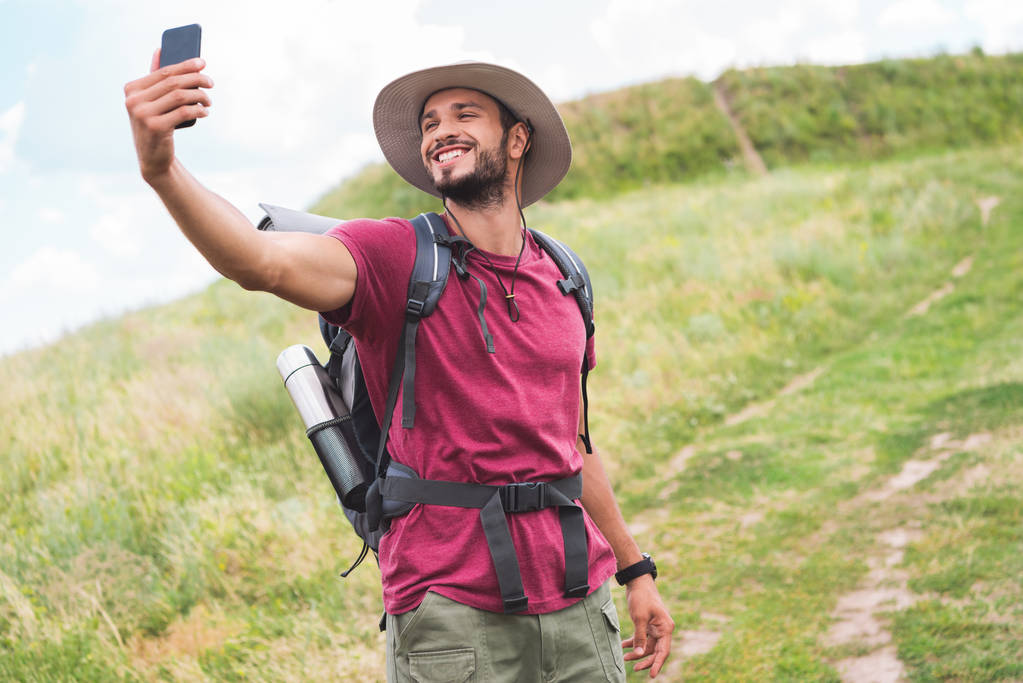 traveler with backpack taking selfie on smartphone on summer meadow - Photo, Image