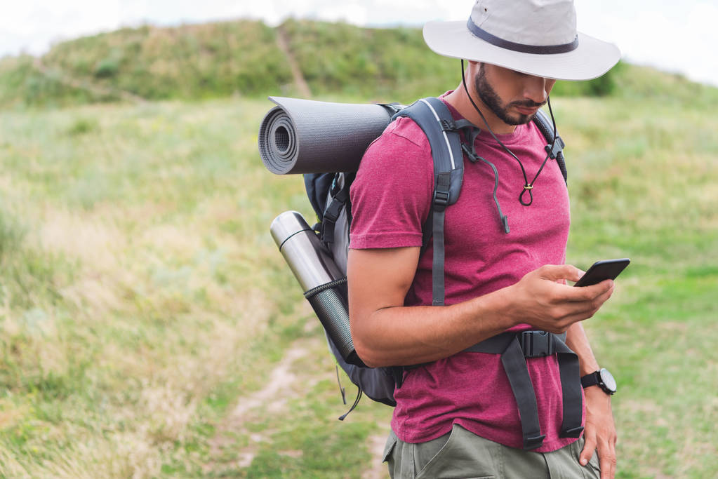 turista en sombrero con mochila usando teléfono inteligente en el prado de verano
 - Foto, imagen