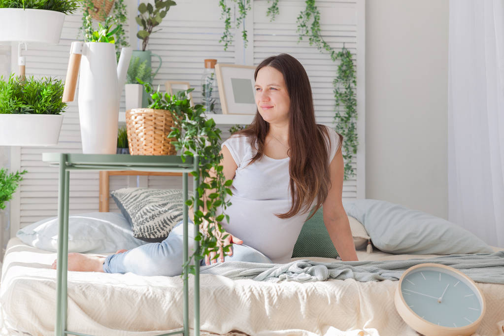 Молодая беременная женщина позирует в светлом современном скандинавском интерьере спальни
 - Фото, изображение