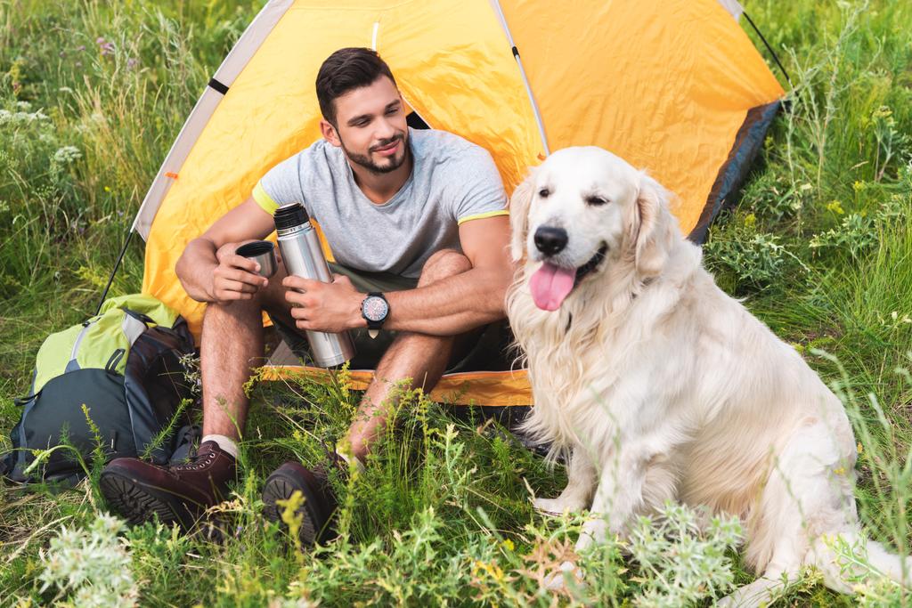 Reisender mit Thermoskanne sitzt im Zelt und schaut Hund an - Foto, Bild
