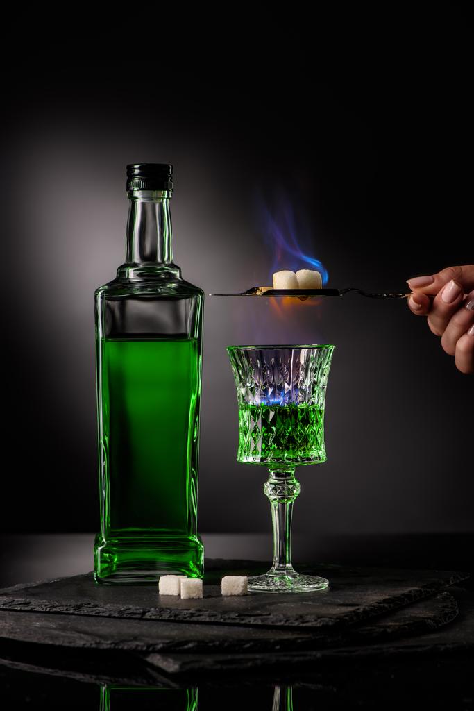 plan recadré de femme tenant cuillère avec des cubes de sucre sur la combustion de l'absinthe dans le verre sur fond sombre
 - Photo, image