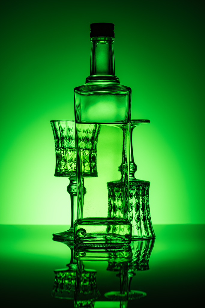 fles absint met lood bril op reflecterende oppervlak en donkere groene achtergrond - Foto, afbeelding