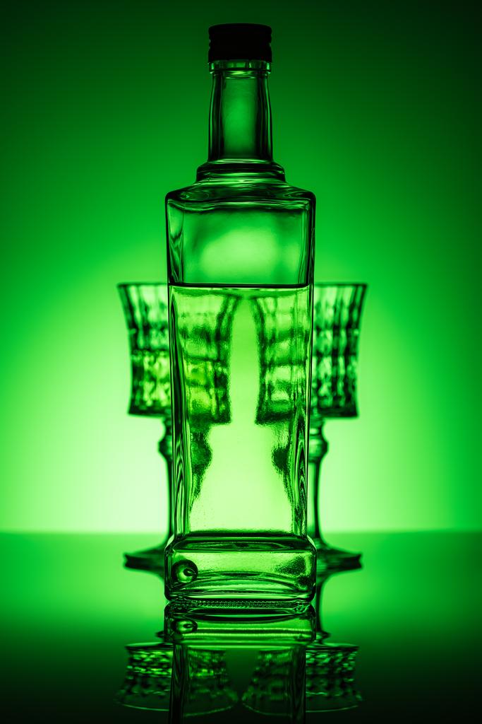 fles absint met bril op spiegelend oppervlak en donker groene achtergrond - Foto, afbeelding