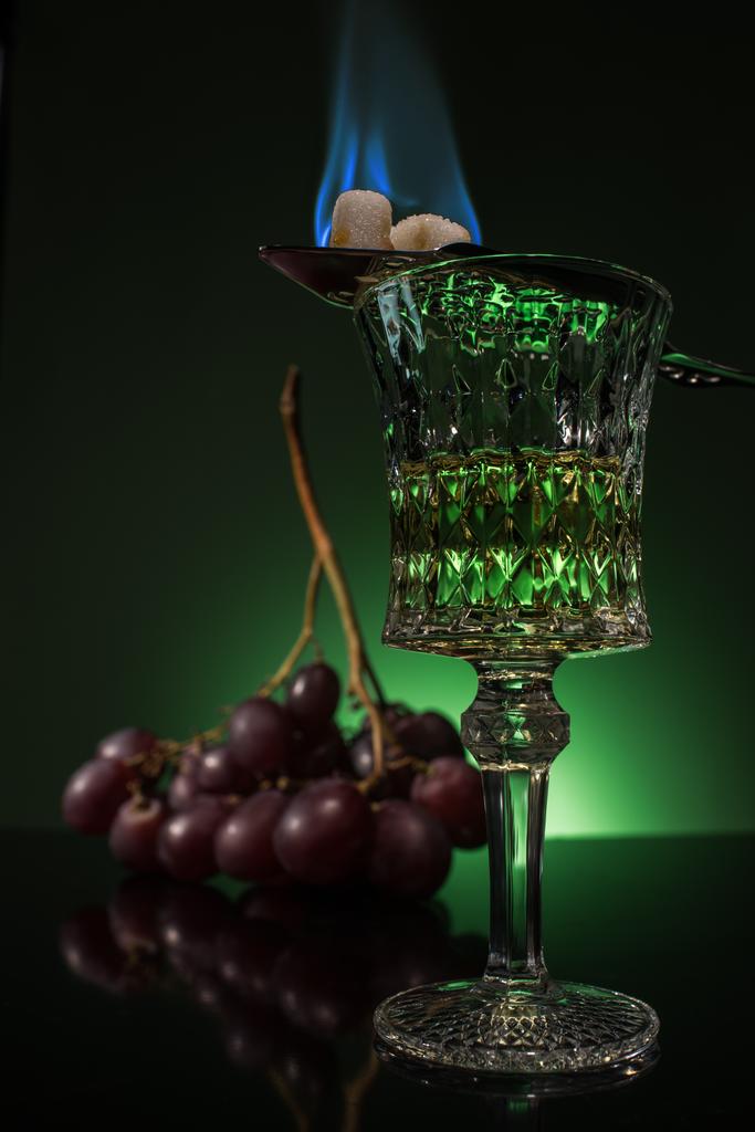 vidro de chumbo de absinto com ramo de uvas e queima de açúcar na colher na superfície reflexiva e fundo verde escuro
 - Foto, Imagem