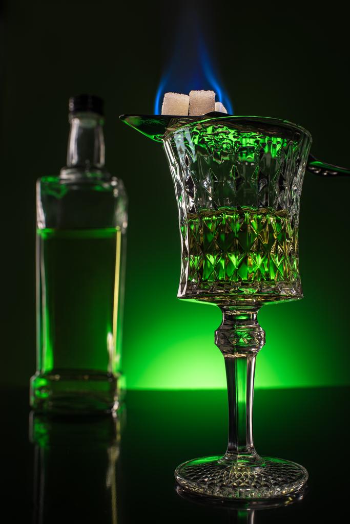 glass and bottle of absinthe with burning sugar on spoon on reflective surface and dark green background - Photo, Image