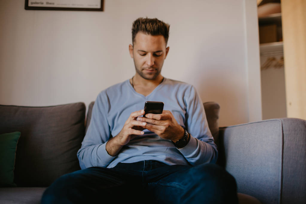 junger gutaussehender Mann, der zu Hause auf dem Sofa sitzt und sein Handy benutzt. Männer halten Smartphone in der Hand und tippen SMS. - Foto, Bild