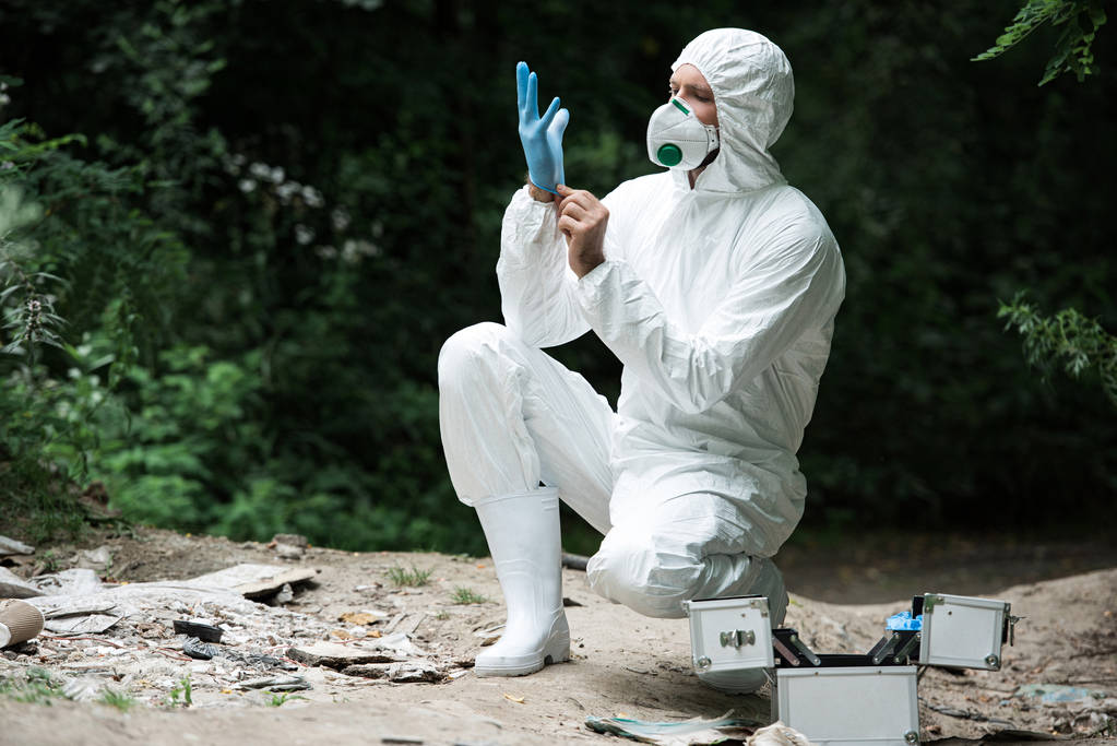 male scientist in protective mask and suit putting on latex glove in forest - Photo, Image